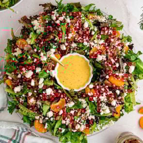 Festive Christmas salad wreath with fresh greens, red pomegranate seeds, white feta crumbles, orange mandarin slices, and pecans around a bowl of creamy dressing.