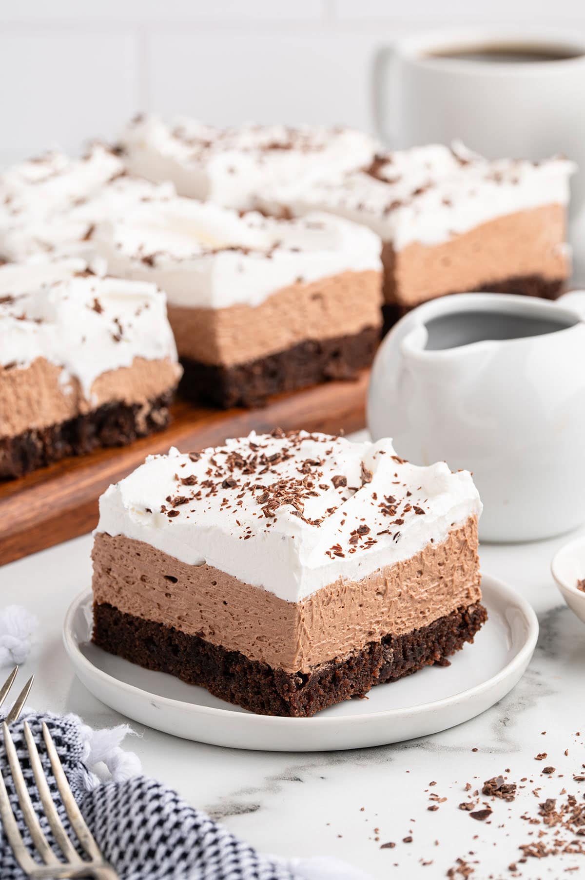 A square slice of French silk brownie sits on a white plate, showing layers of fudgy chocolate base, smooth mousse, and whipped cream topping.