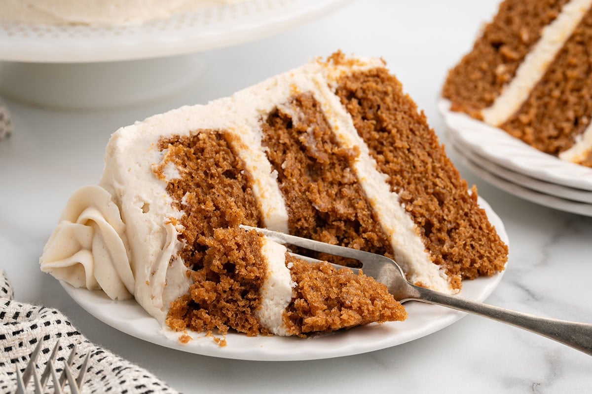 A slice of gingerbread cake topped with creamy white frosting sits on a white plate.