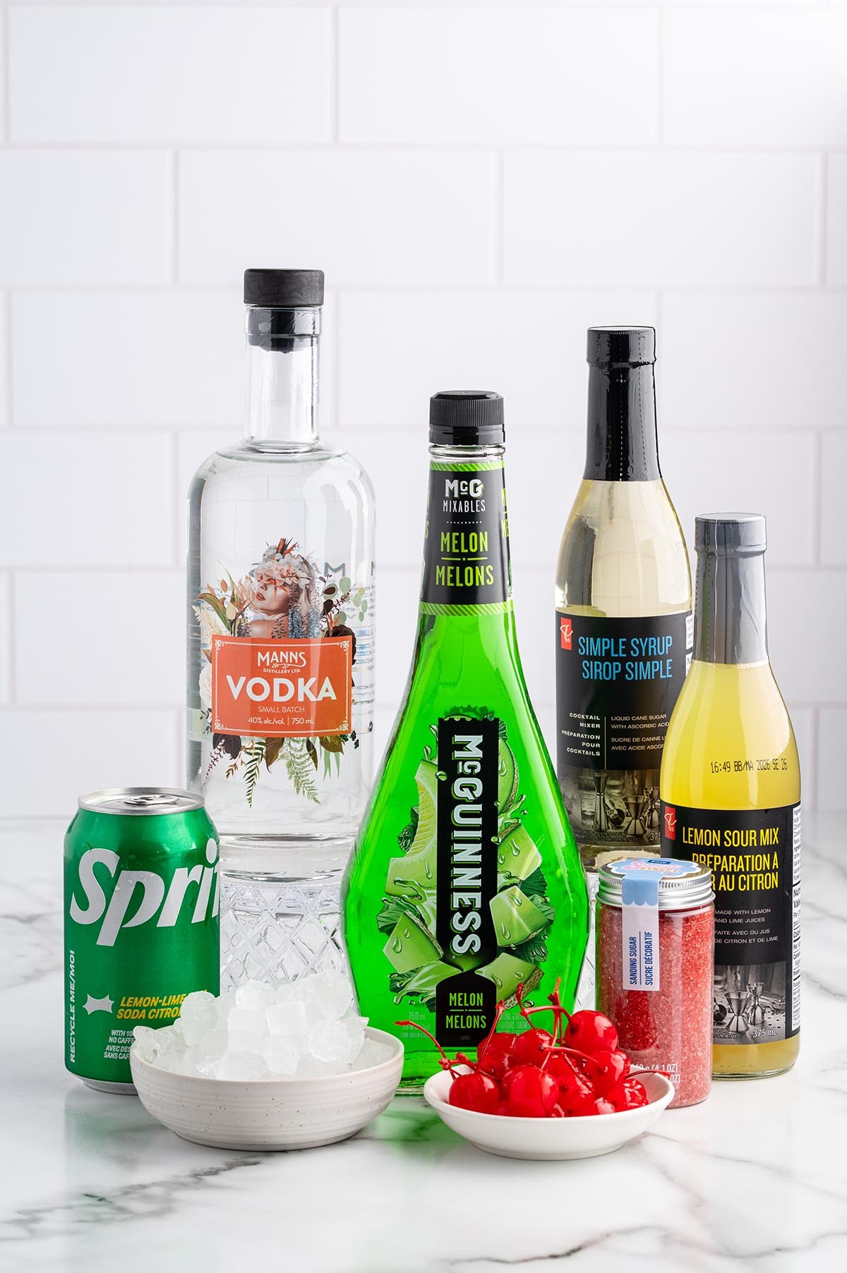 Ingredients for a Grinch cocktail sit on a marble counter: melon vodka bottle, simple syrup, Sprite can, ice bowl, and cherries.