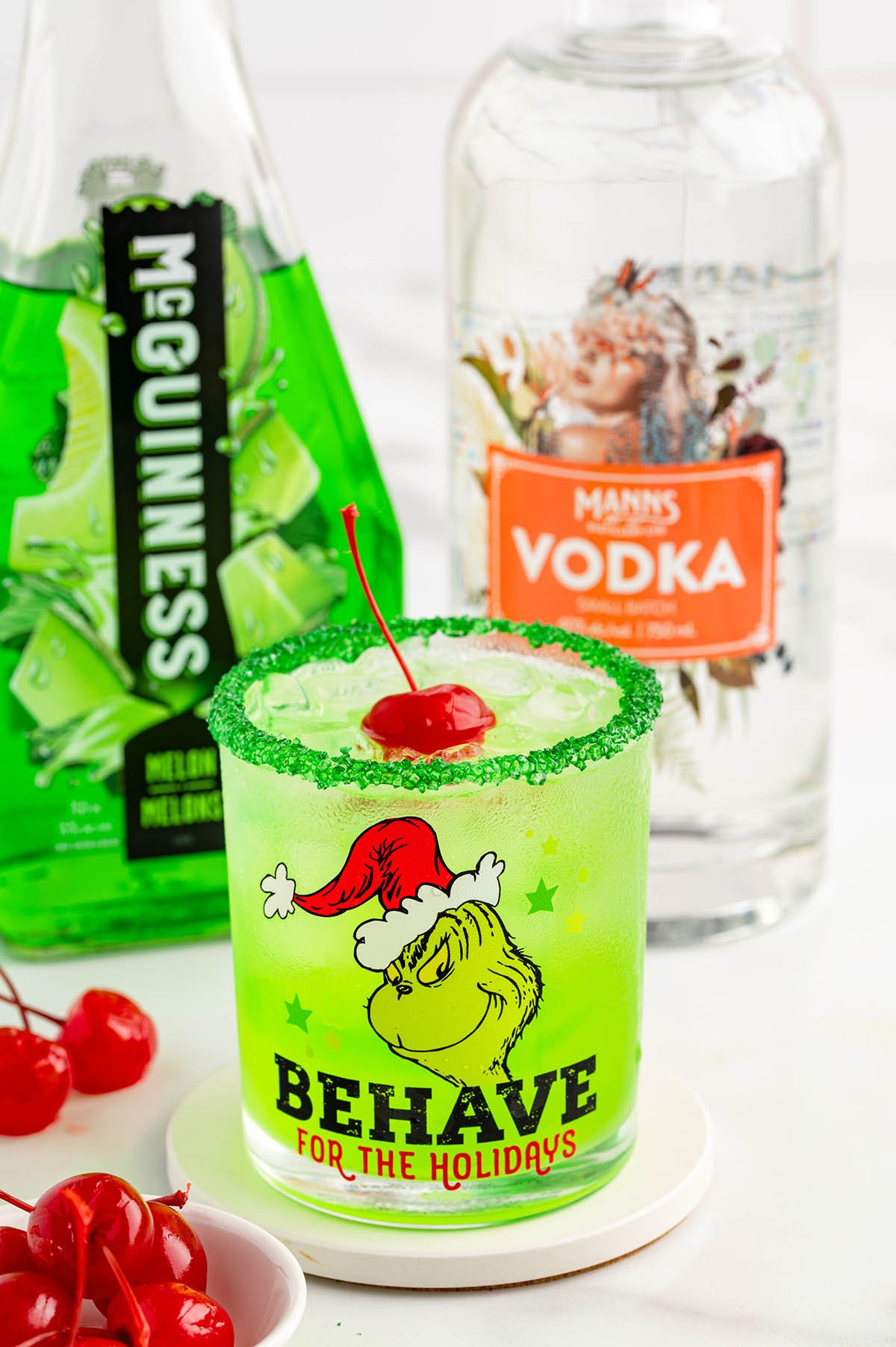 A festive Grinch cocktail in a green glass with a sugar rim and cherry garnish sits next to holiday-themed vodka and beer bottles.