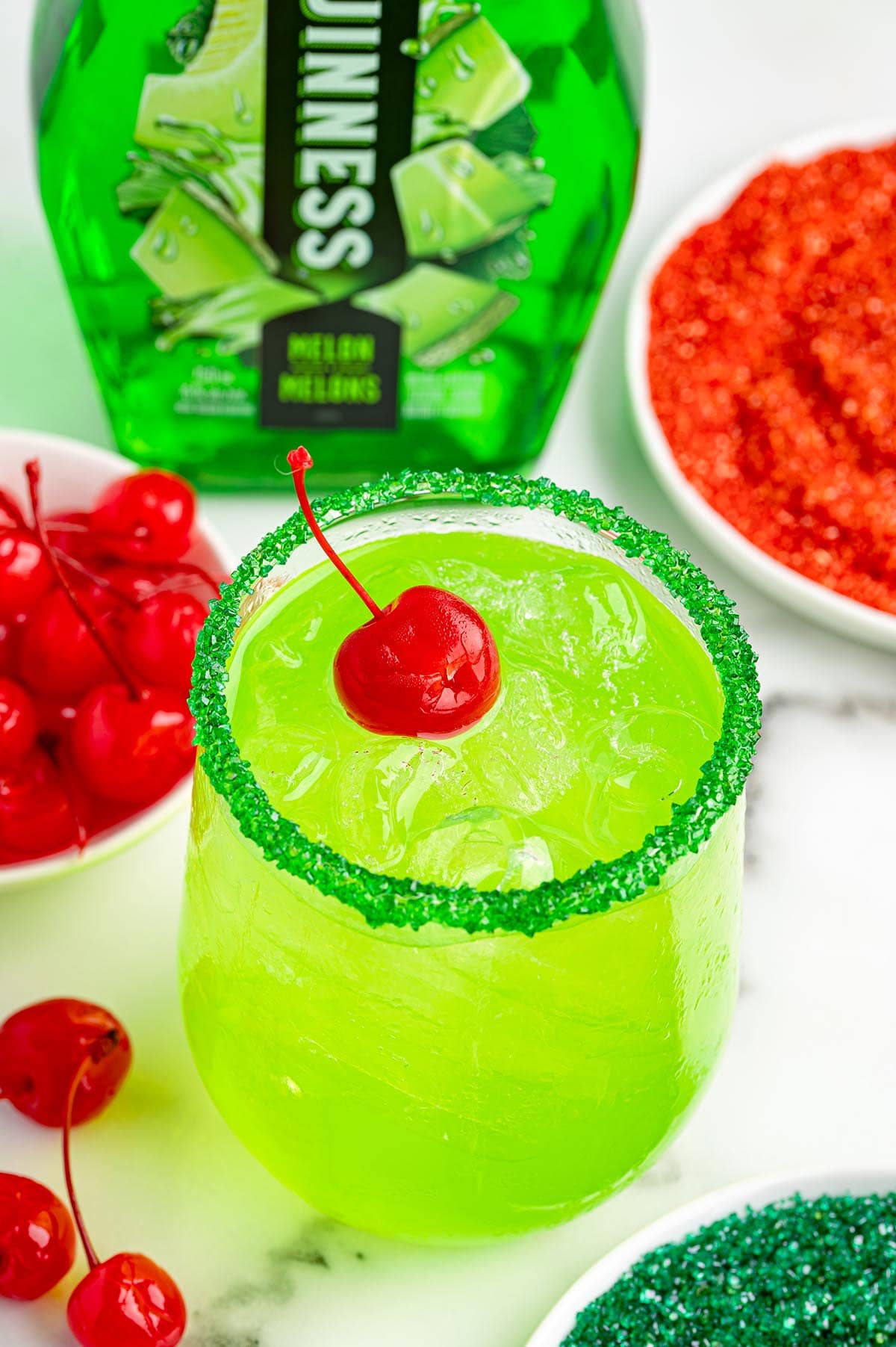 A vibrant green Grinch Cocktail sits in a glass with a green sugar rim and a cherry garnish.