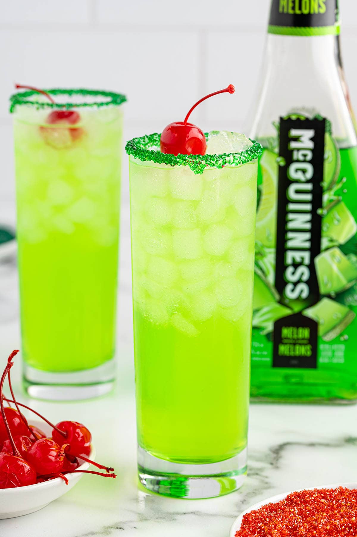 Two tall glasses of green Grinch cocktails sit on a white counter with a bottle of McGuinness Melon Liqueur nearby.