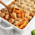 Wooden spoon scooping cheesy meatball penne casserole from a white baking dish.