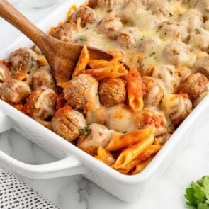 Wooden spoon scooping cheesy meatball penne casserole from a white baking dish.