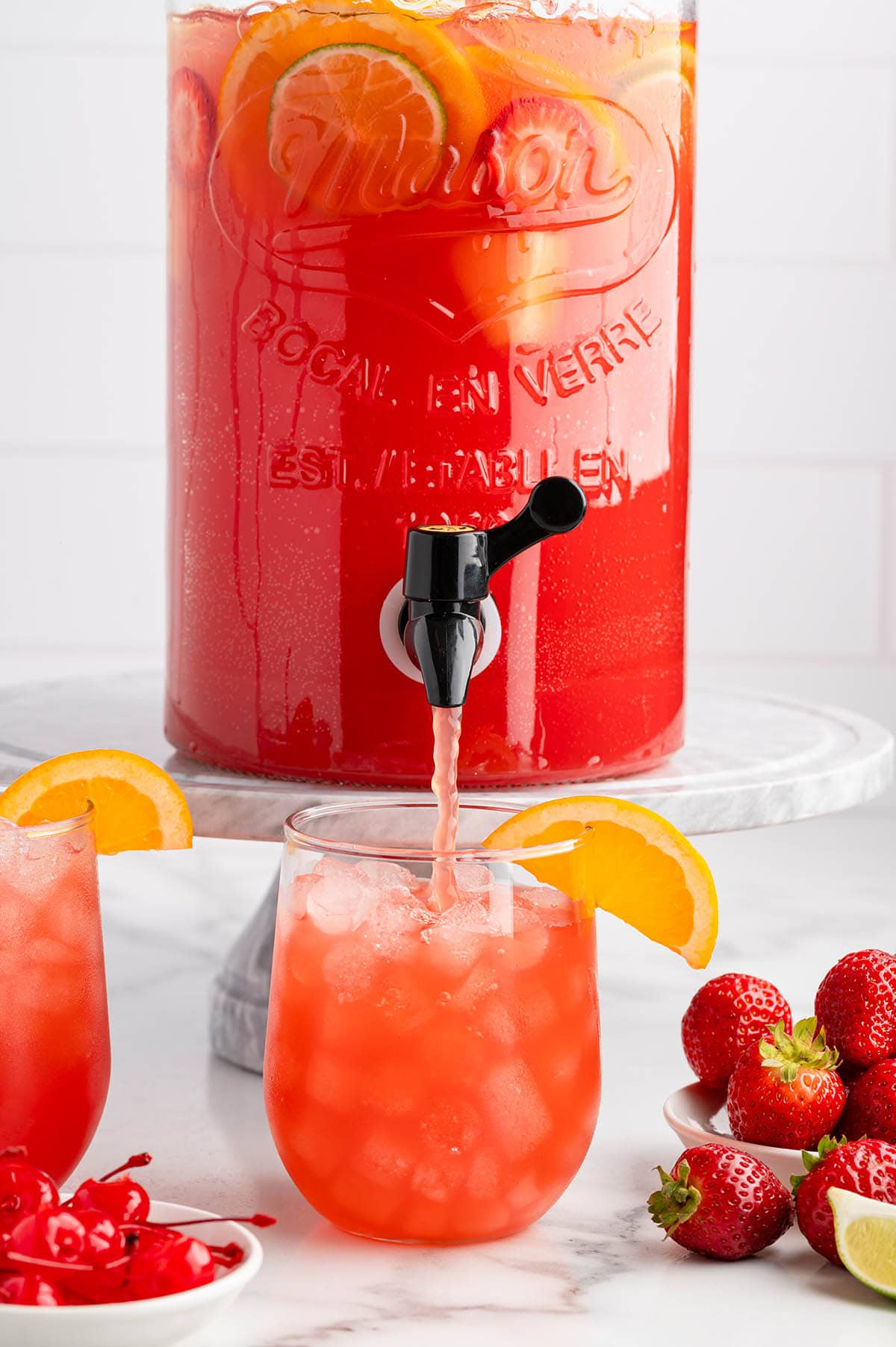 Dispenser of party punch pouring punch into a glass with ice and an orange garnish