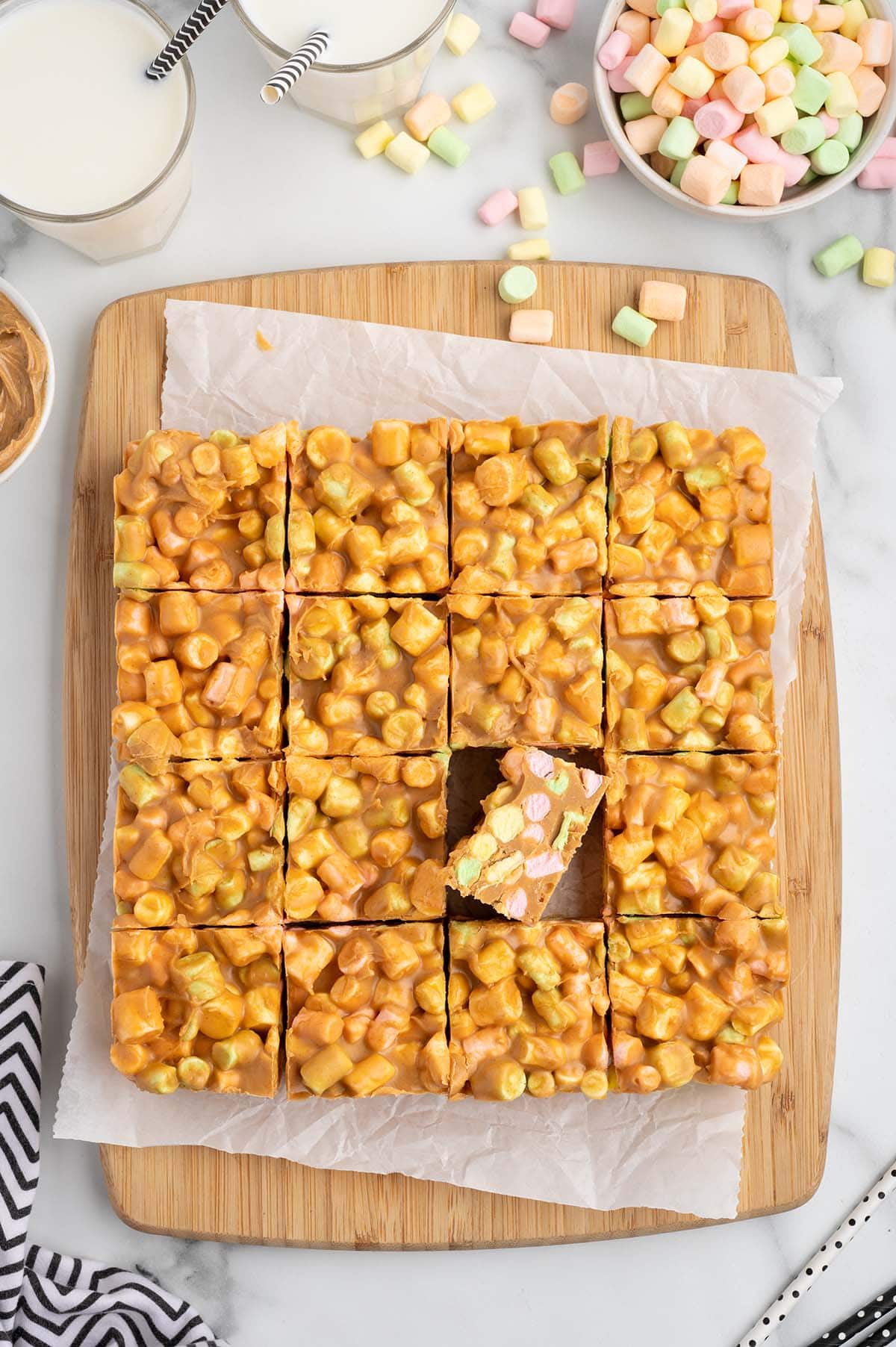 Golden peanut butter marshmallow squares cut into pieces on a wooden board, next to glasses of milk and a bowl of colorful marshmallows.