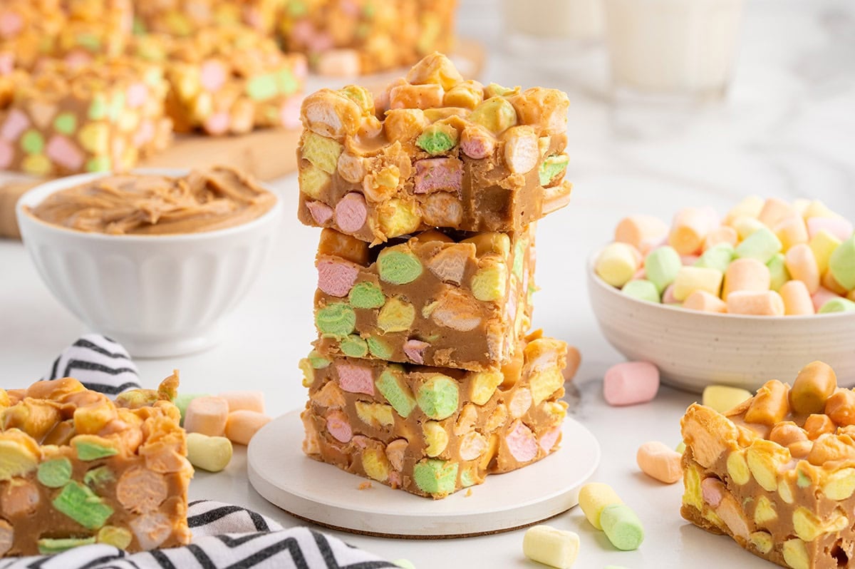 A stack of peanut butter marshmallow squares topped with colorful cereal bits sits next to a bowl of peanut butter and a glass of milk.