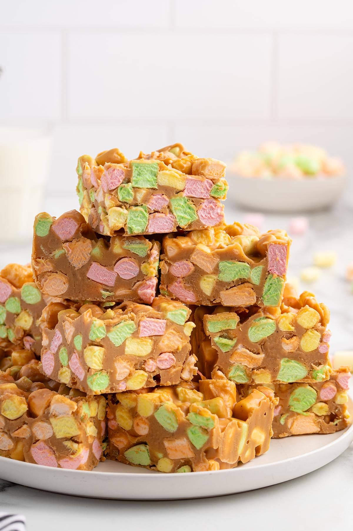 Stack of colorful peanut butter marshmallow squares on a white plate next to a glass of milk.