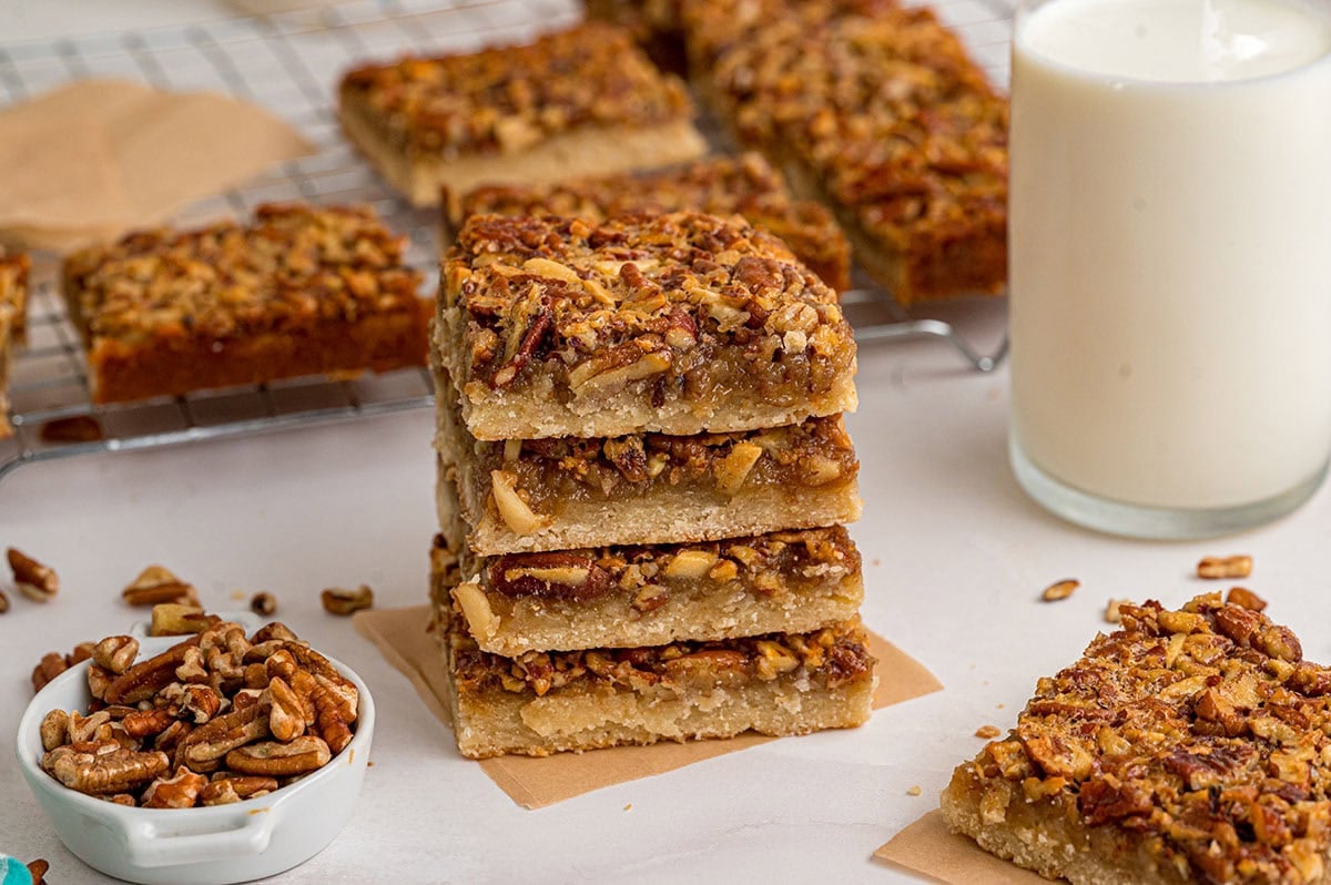 Stack of golden pecan pie bars cooling on a wire rack next to a glass of milk.