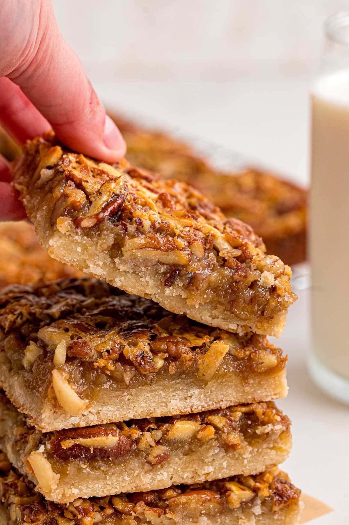 A hand lifts a stack of four pecan pie bars from the pan, topped with nuts and syrup, beside a glass of milk.