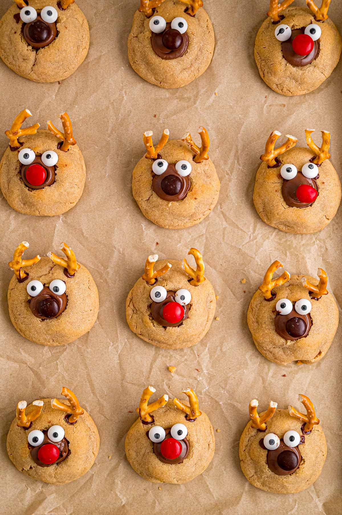 Reindeer peanut butter blossom cookies with pretzel antlers and red noses sit on parchment paper.