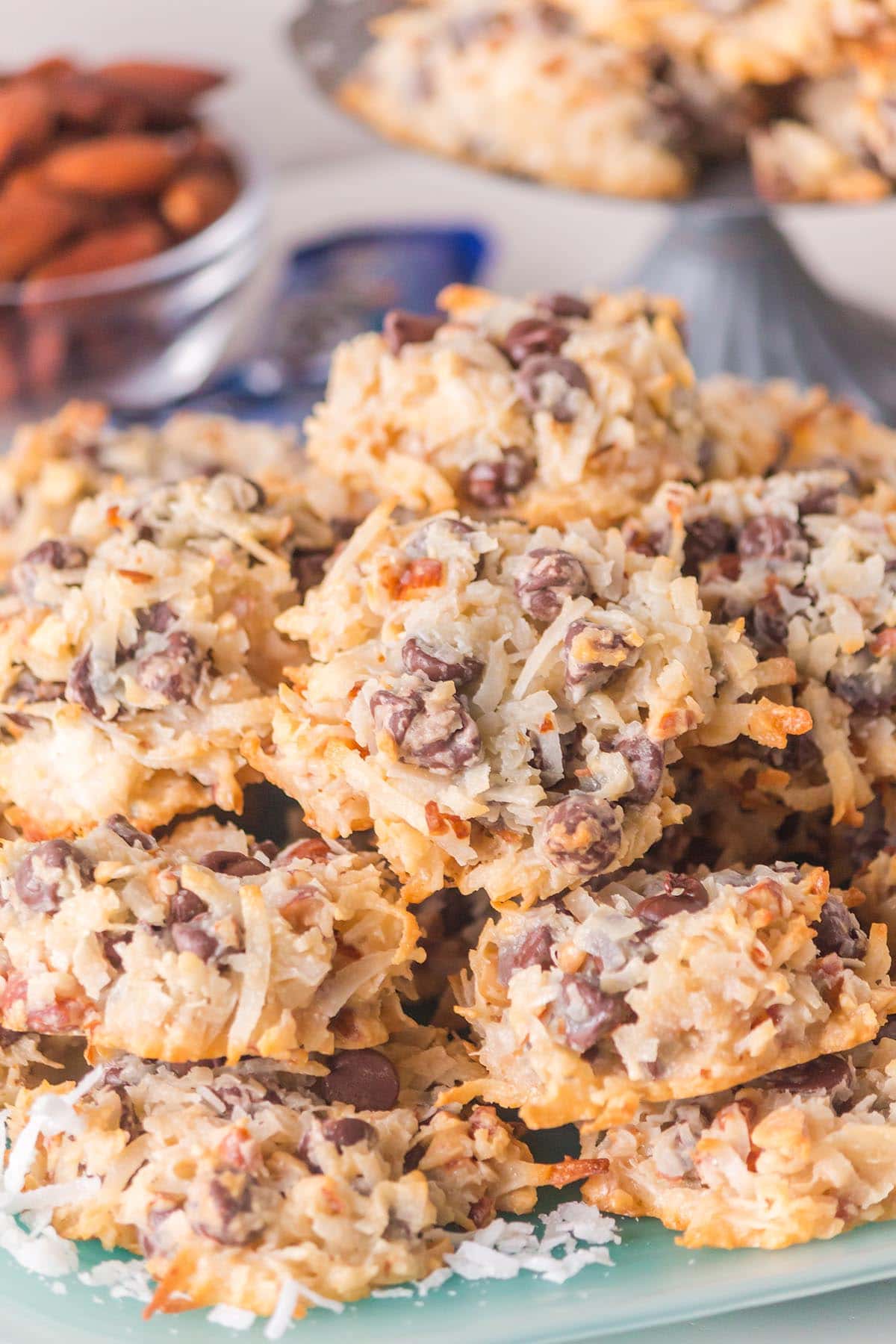 A stack of golden Almond Joy cookies topped with coconut shreds, chocolate chips, and almond pieces on a clear plate.