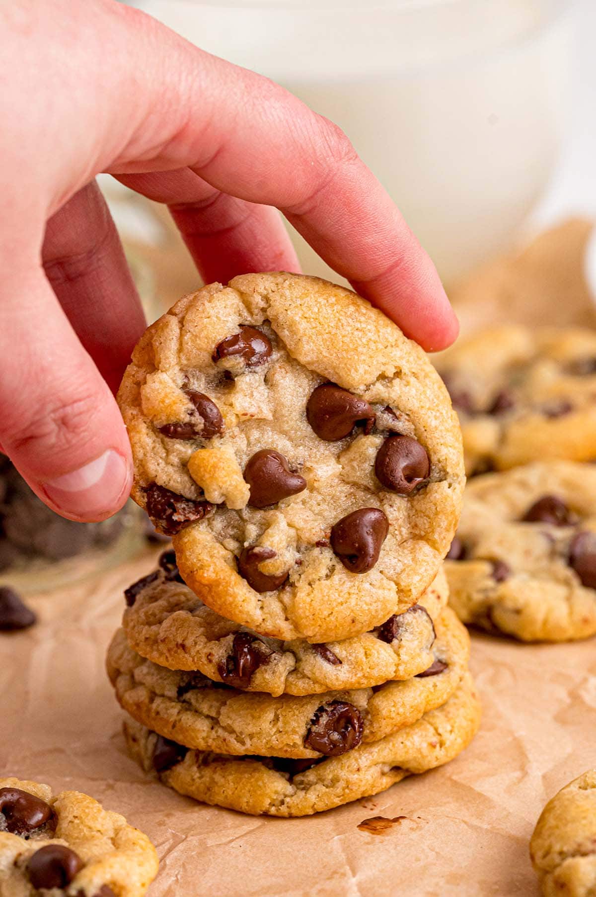 A hand holds a golden eggless chocolate chip cookie over a stack of fresh baked ones on parchment paper with milk nearby.