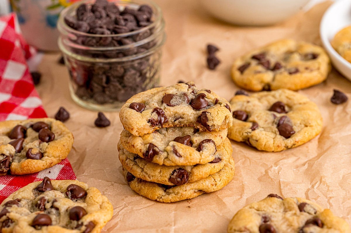 Freshly baked eggless chocolate chip cookies stacked on parchment paper with chocolate chips nearby.