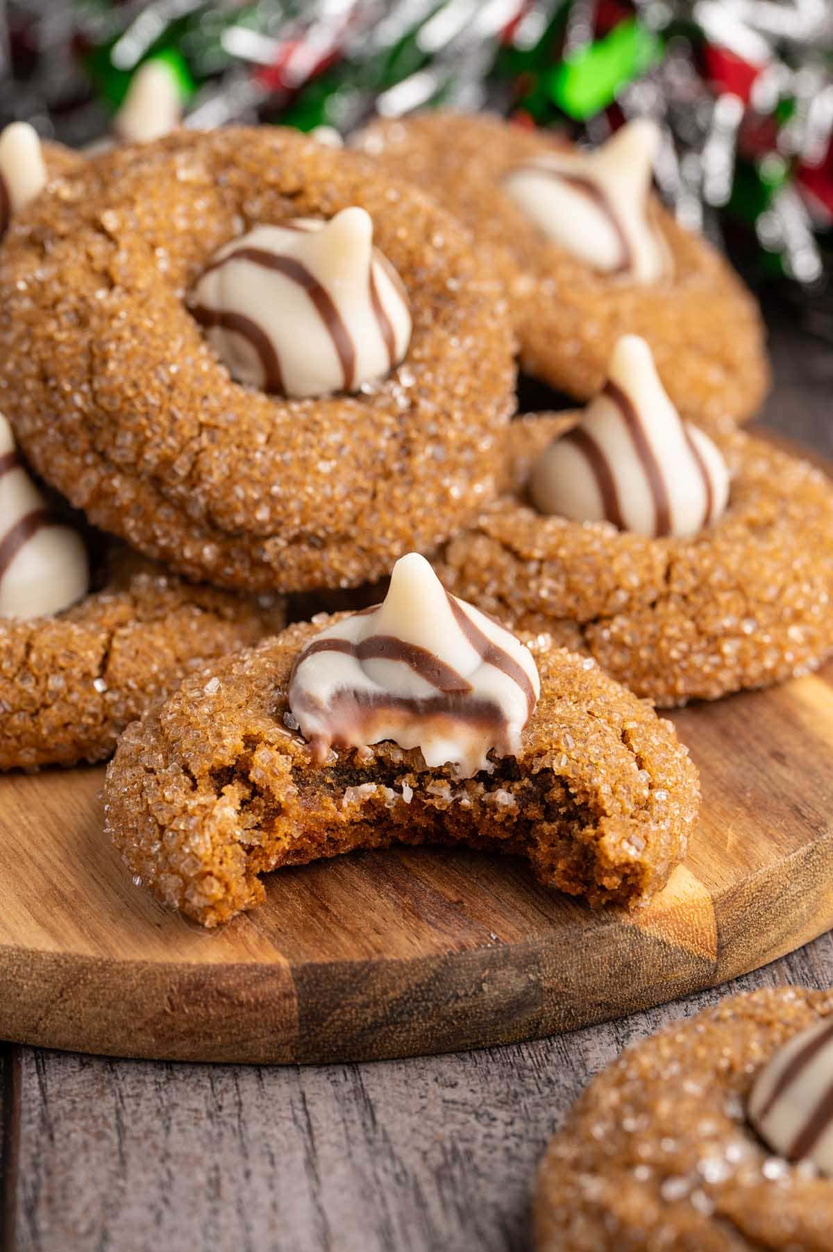 A stack of soft gingerbread cookies topped with chocolate kisses sits on a wooden board, with one cookie bitten to show the gooey center.
