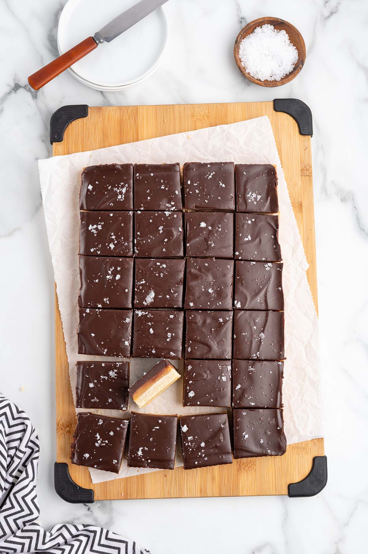 Fresh millionaire shortbread bars topped with dark chocolate and sea salt sit on a wooden cutting board.