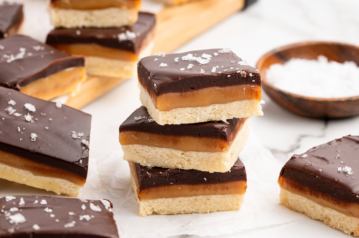 Stacked millionaire shortbread bars with chocolate tops and sea salt flakes on a wooden board.