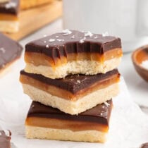 Stacked millionaire shortbread bars show a shortbread base, gooey caramel middle, and dark chocolate top sprinkled with sea salt flakes.