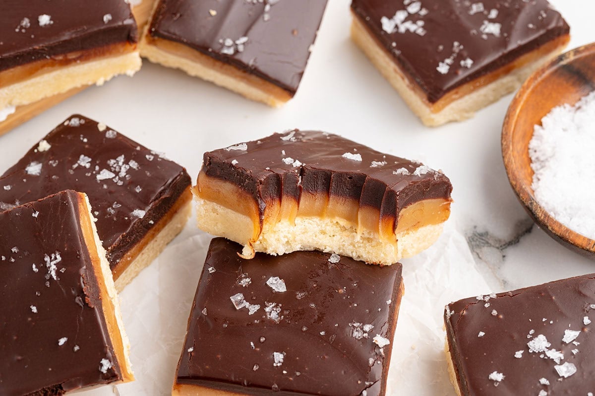 Millionaire shortbread bars with chocolate tops and sea salt flakes sit on a white surface.