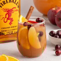 Fireball sangria glass with apple slices, orange wedges, cinnamon stick, and cherries sits by the whisky bottle.