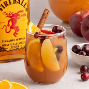 Fireball sangria glass with apple slices, orange wedges, cinnamon stick, and cherries sits by the whisky bottle.