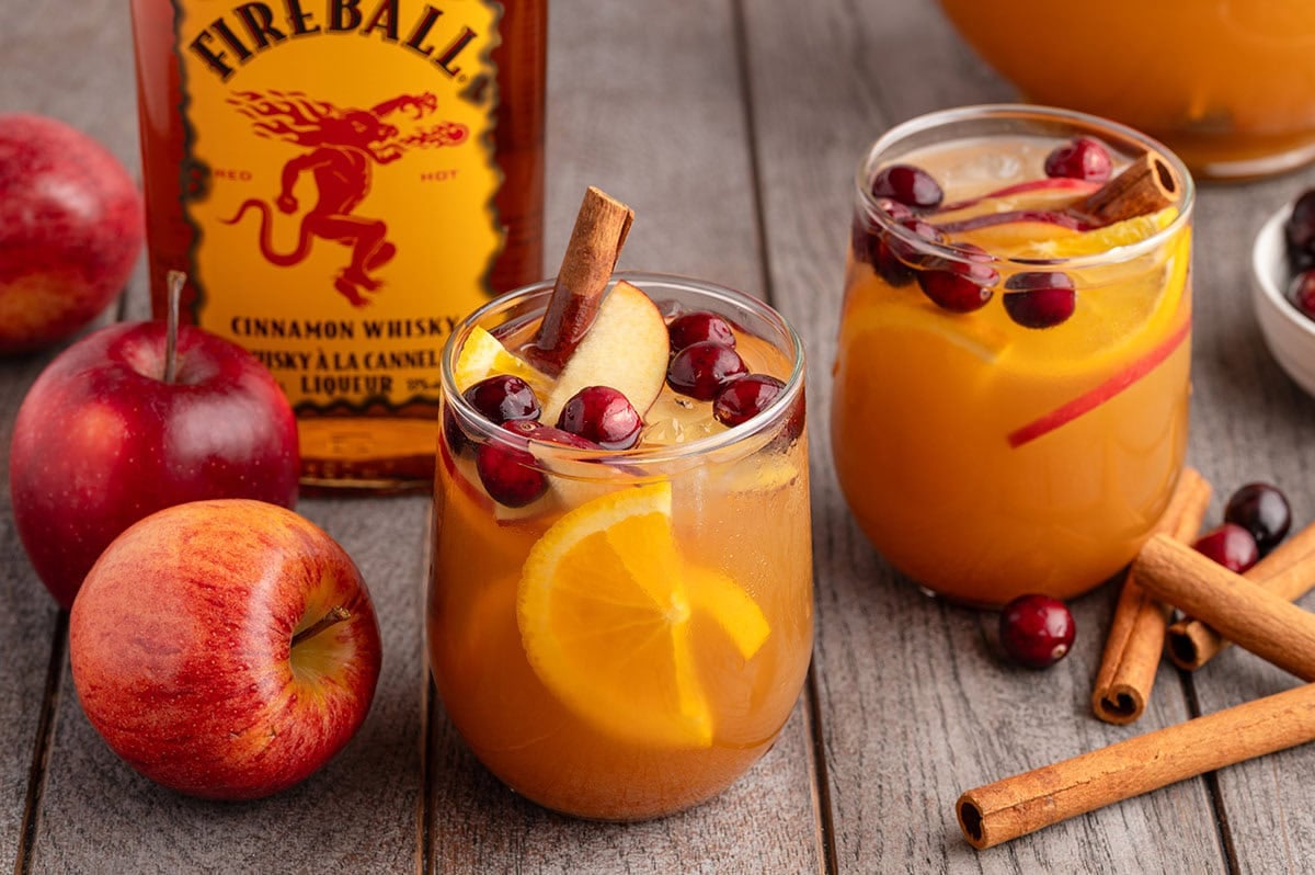 Two glasses of Fireball sangria with cranberries, apple slices, orange wedges, and cinnamon sticks sit next to the whisky bottle.
