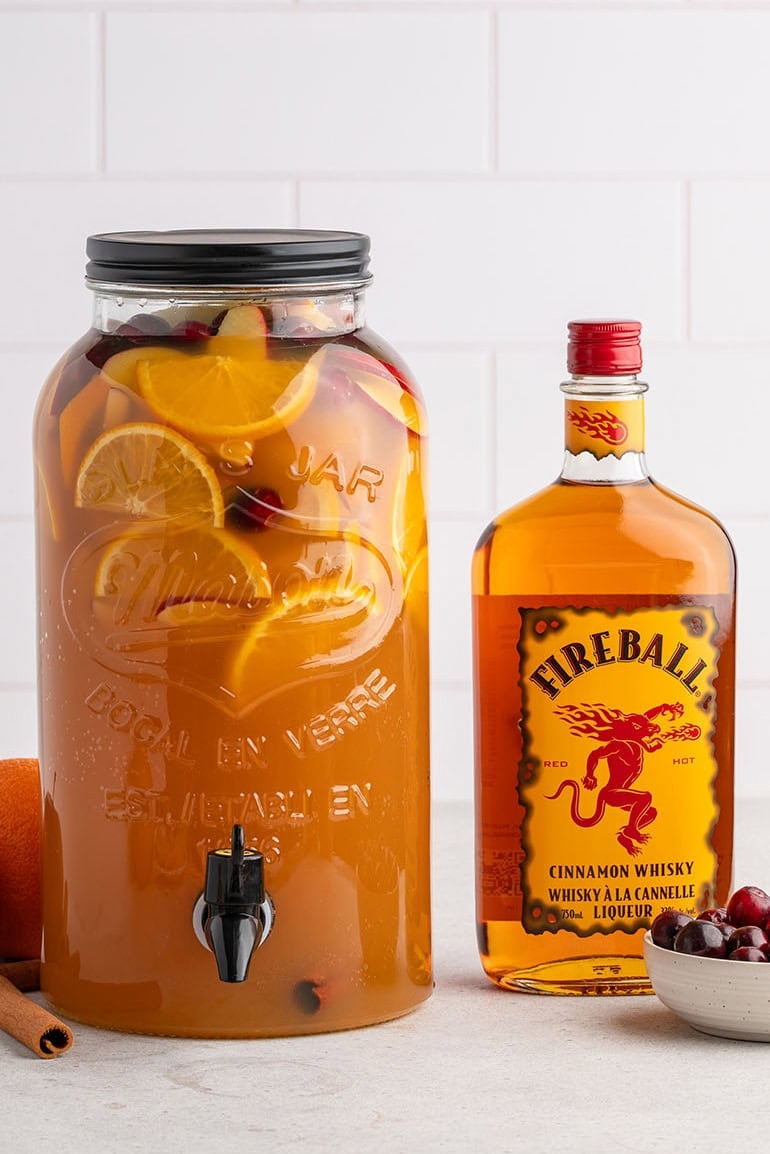 Glass dispenser holds Fireball sangria with orange slices, apple chunks, cranberries, and cinnamon sticks next to the whisky bottle.