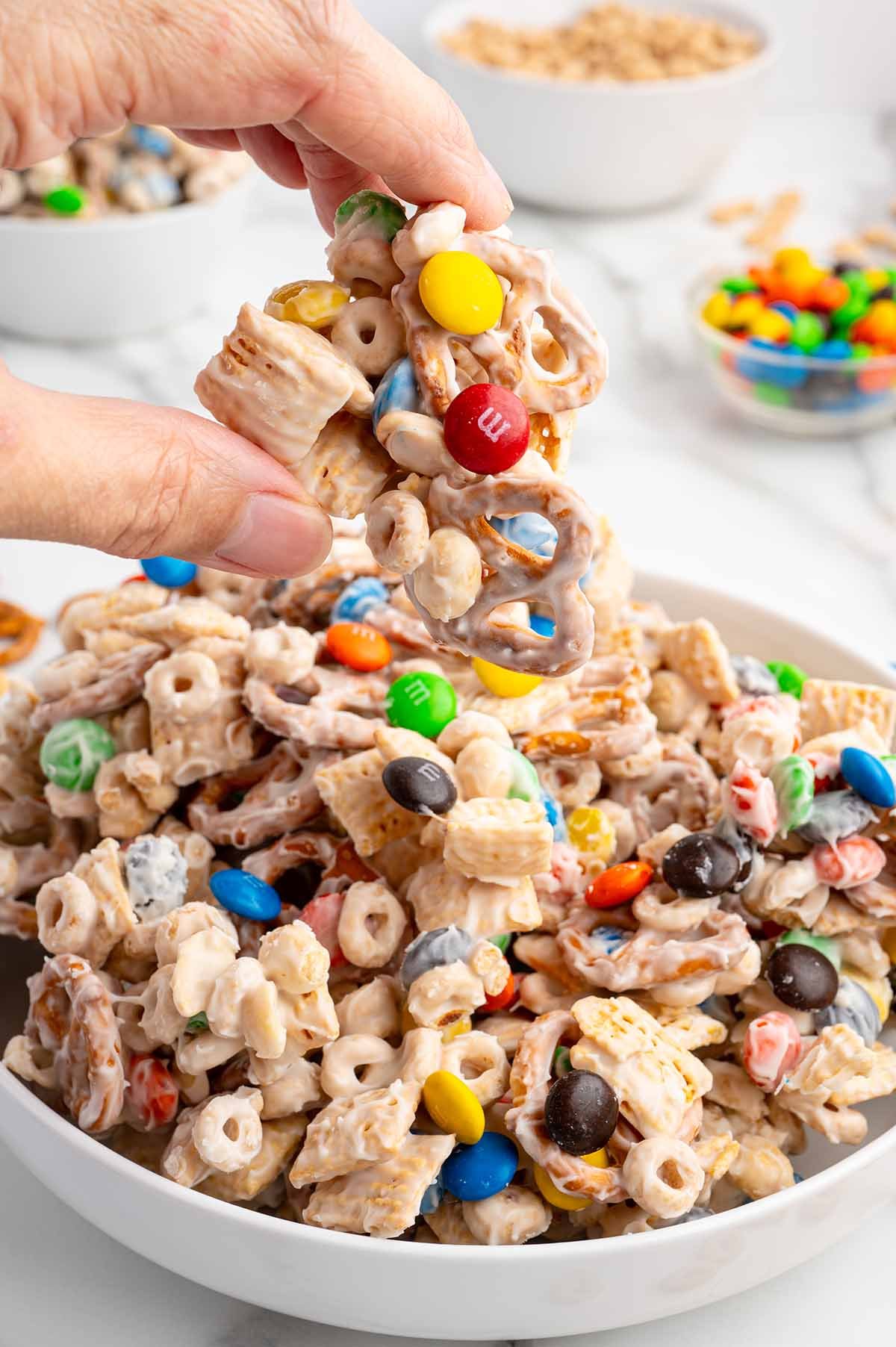 A hand lifts a piece of white chocolate-covered snack mix from a big bowl full of pretzels, cereal, and colorful M&Ms.