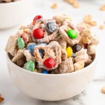 A white bowl holds a mix of white-coated Chex cereal, colorful M&Ms, pretzels, and peanuts.