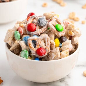 A white bowl holds a mix of white-coated Chex cereal, colorful M&Ms, pretzels, and peanuts.