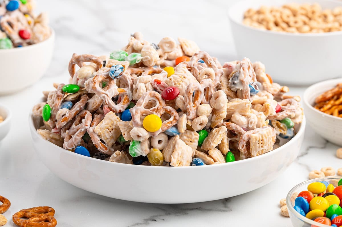 A big bowl of white trash snack mix sits on a white counter, full of white chocolate coated Chex, pretzels, and colorful M&Ms.