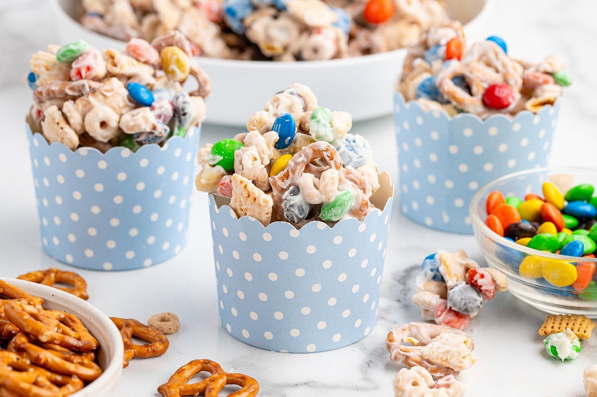 Colorful White Trash snack mix in blue polka-dot cups, with pretzels and M&Ms scattered around.