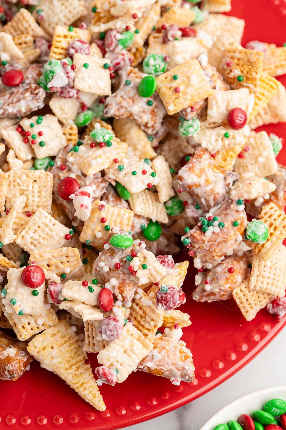 Christmas Chex Mix on a red plate with white chocolate, red M&Ms, and green candies.