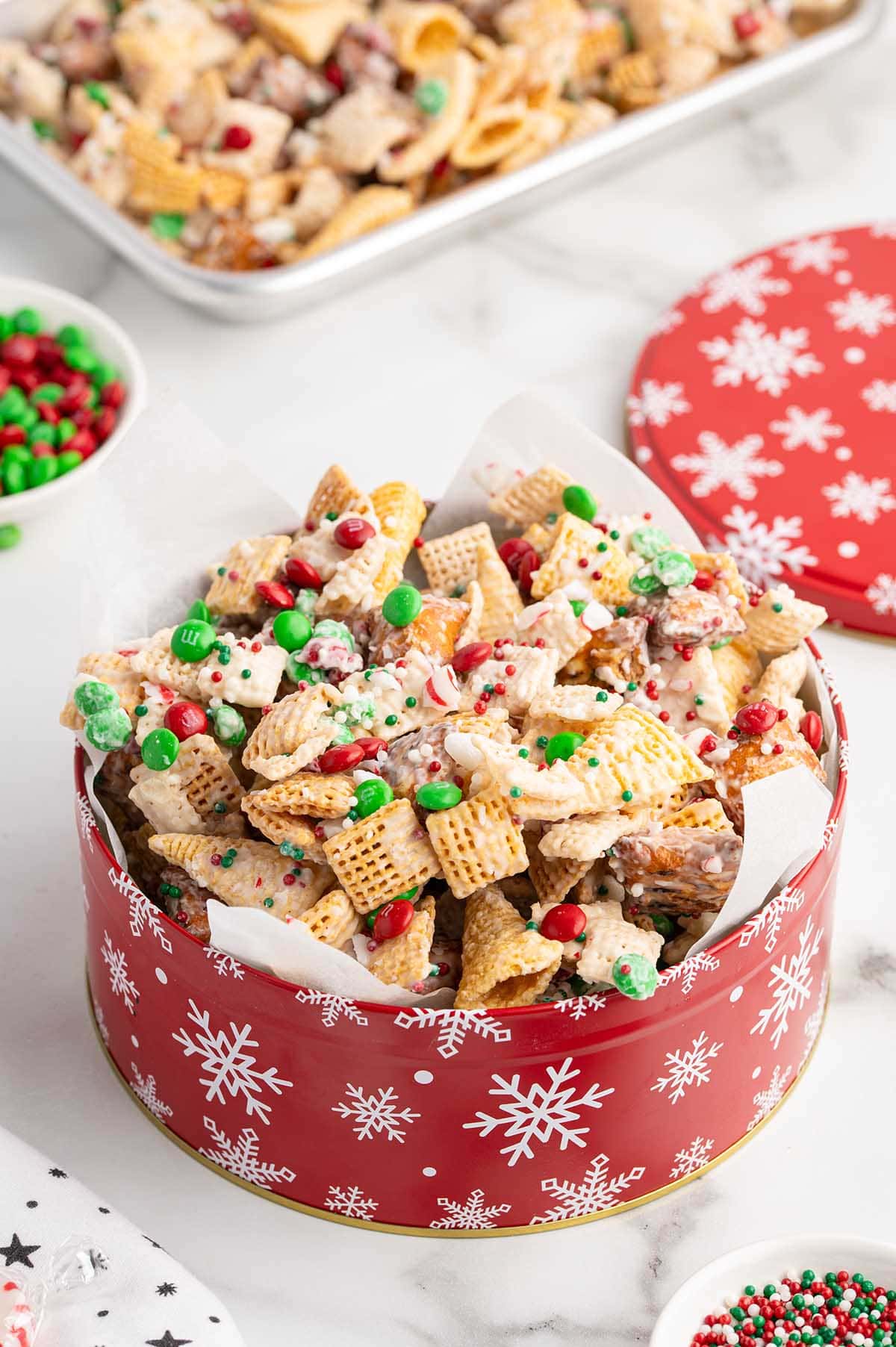 Christmas Chex Mix fills a red snowflake tin with green and red candies on top.