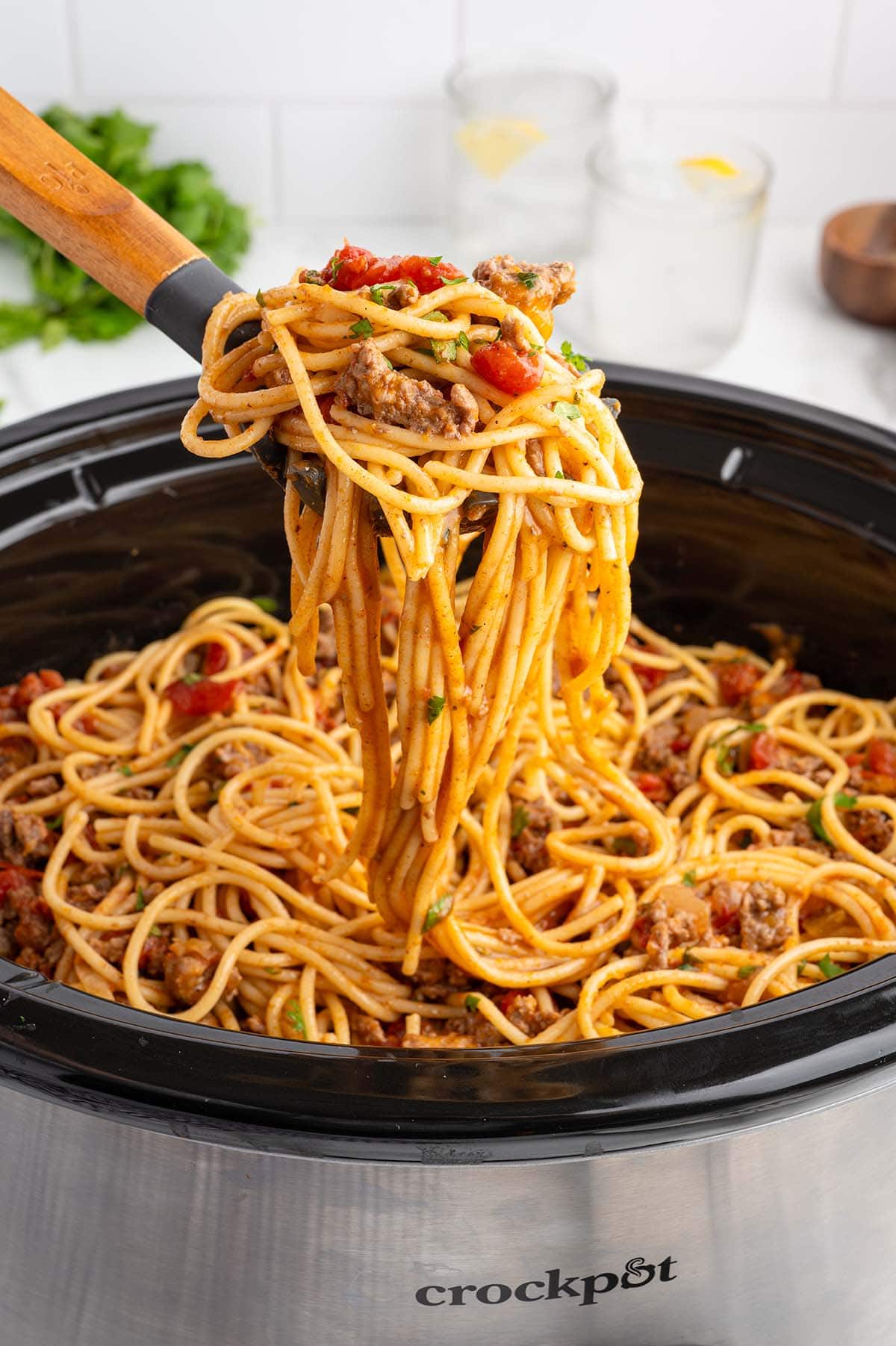 Crockpot taco spaghetti cooks in a slow cooker, wooden spoon lifts saucy noodles with beef and tomatoes.