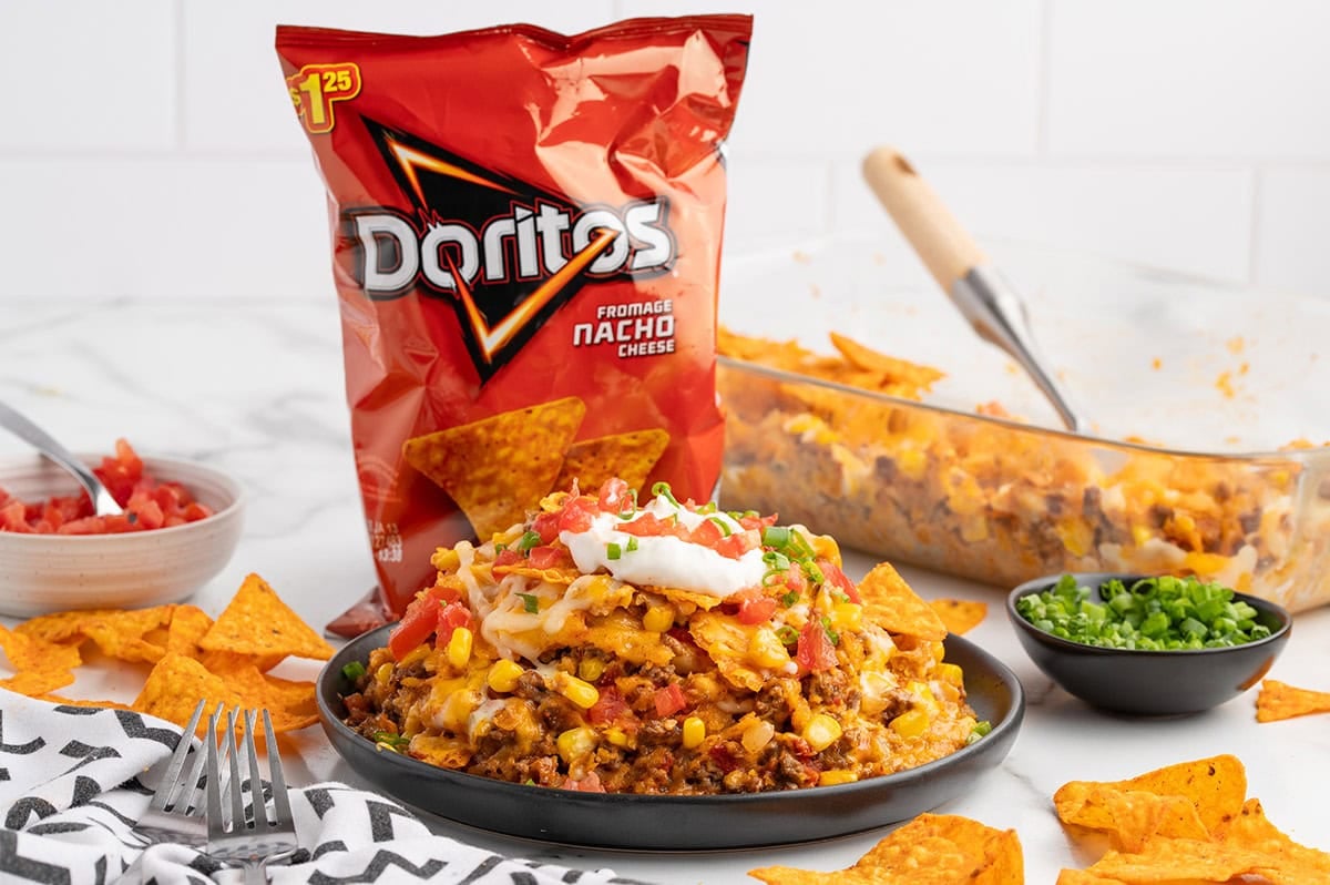 Dorito casserole on a black plate topped with cheese, beef, tomatoes, sour cream, and green onions next to a bag of Flamin' Hot Nacho Doritos.
