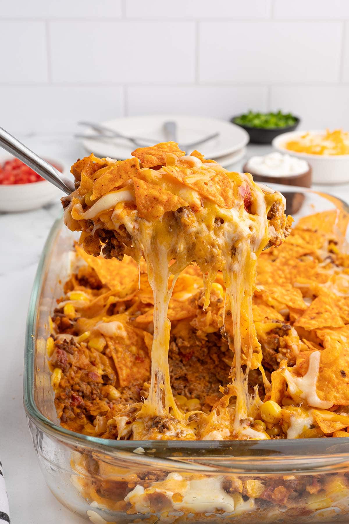 A spoon scoops cheesy Dorito casserole from a glass dish, with cheese stretching long.