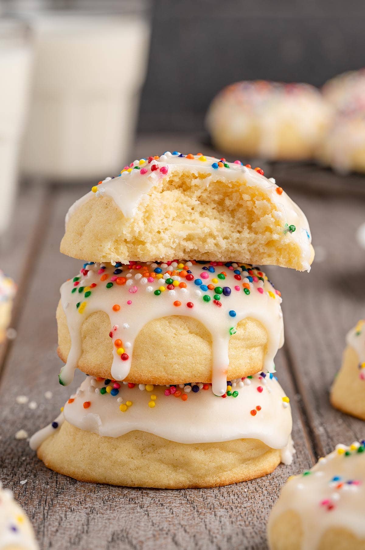Stack of three Italian cookies glazed with white icing and rainbow sprinkles, one bitten open to show soft inside, milk glass nearby.