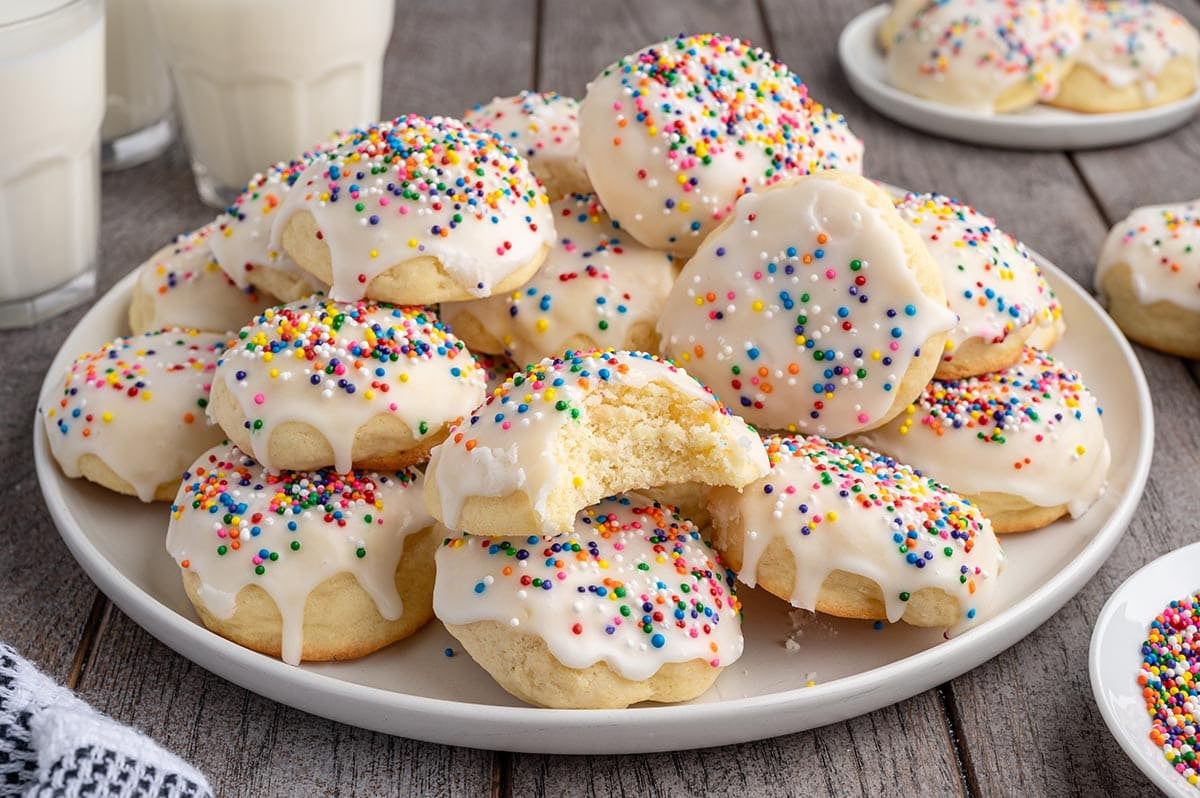 Plate of soft Italian cookies topped with colorful sprinkles and white glaze, served with milk glasses on wood.