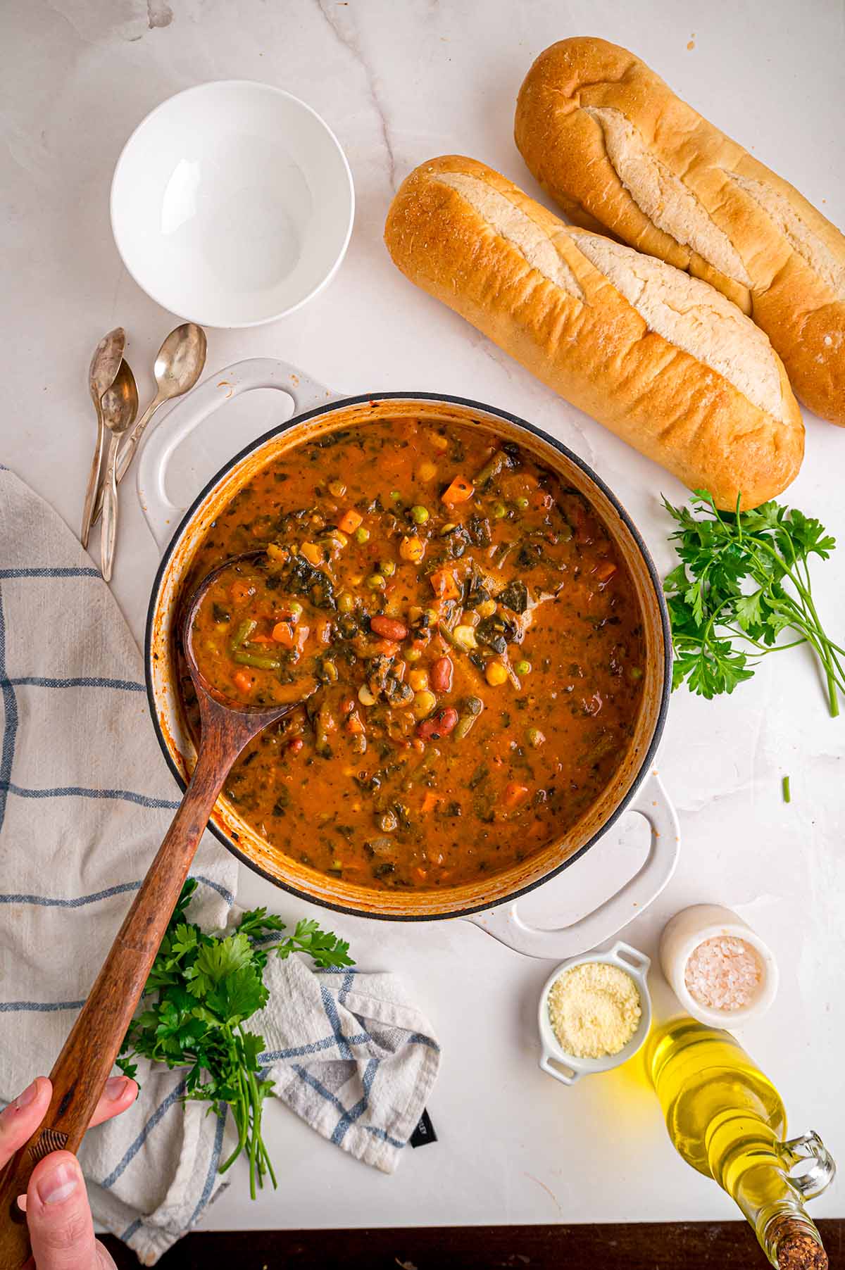 Pot of minestrone soup with beans and veggies, wooden spoon inside, baguettes and parsley on the side.