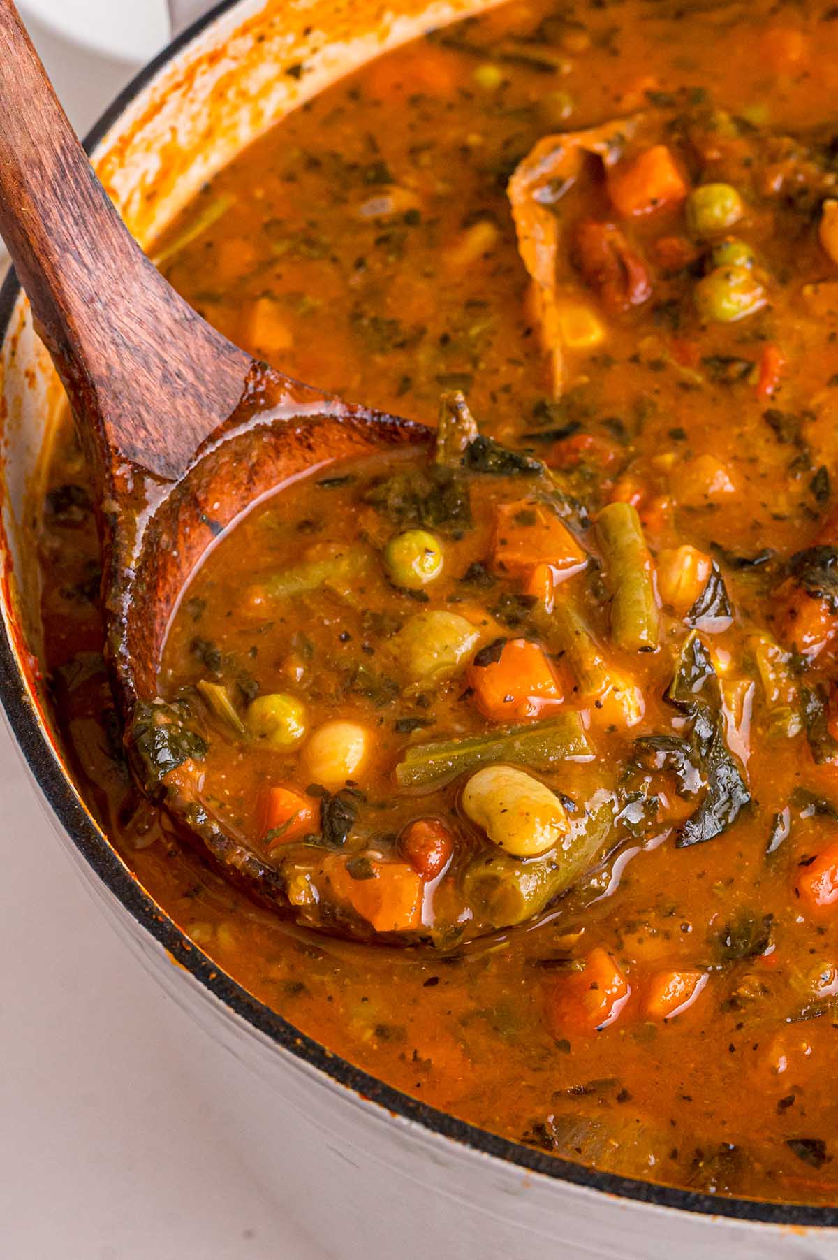 Wooden spoon scoops minestrone soup from a pot full of carrots, green beans, peas, and chickpeas.