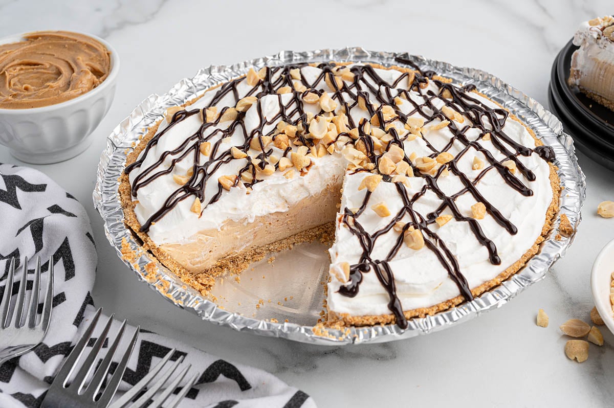 Slice of no-bake peanut butter pie with whipped cream, chocolate drizzle, and peanuts on foil plate.