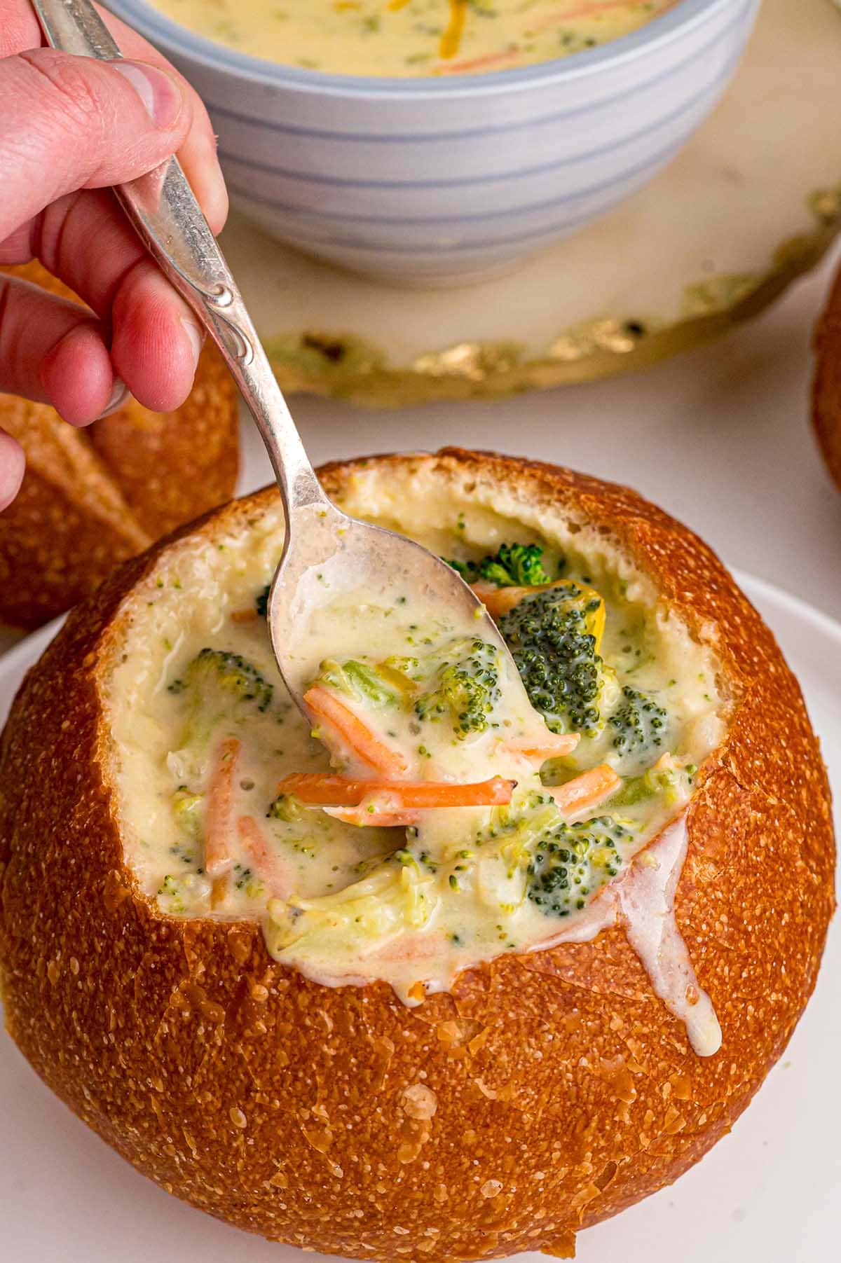 Hand scoops creamy broccoli cheddar soup from a round bread bowl on a white plate.