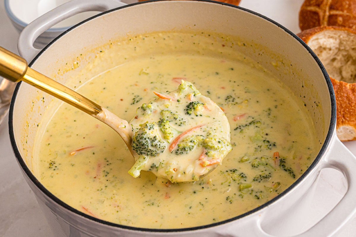 Creamy Panera broccoli cheddar soup in a white pot with a gold ladle scooping broccoli and carrots.