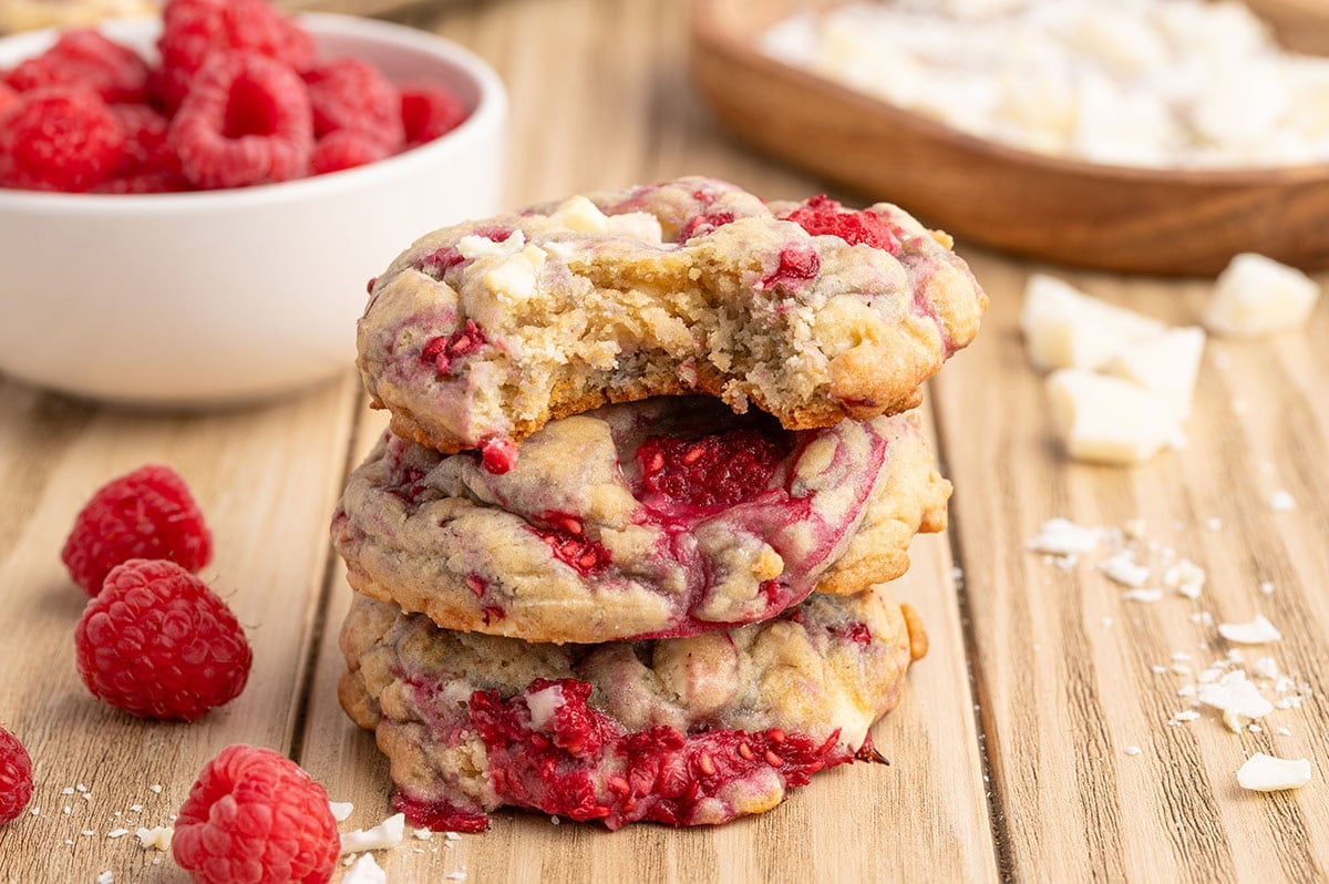 Stack of three white chocolate raspberry cookies sits next to fresh raspberries.