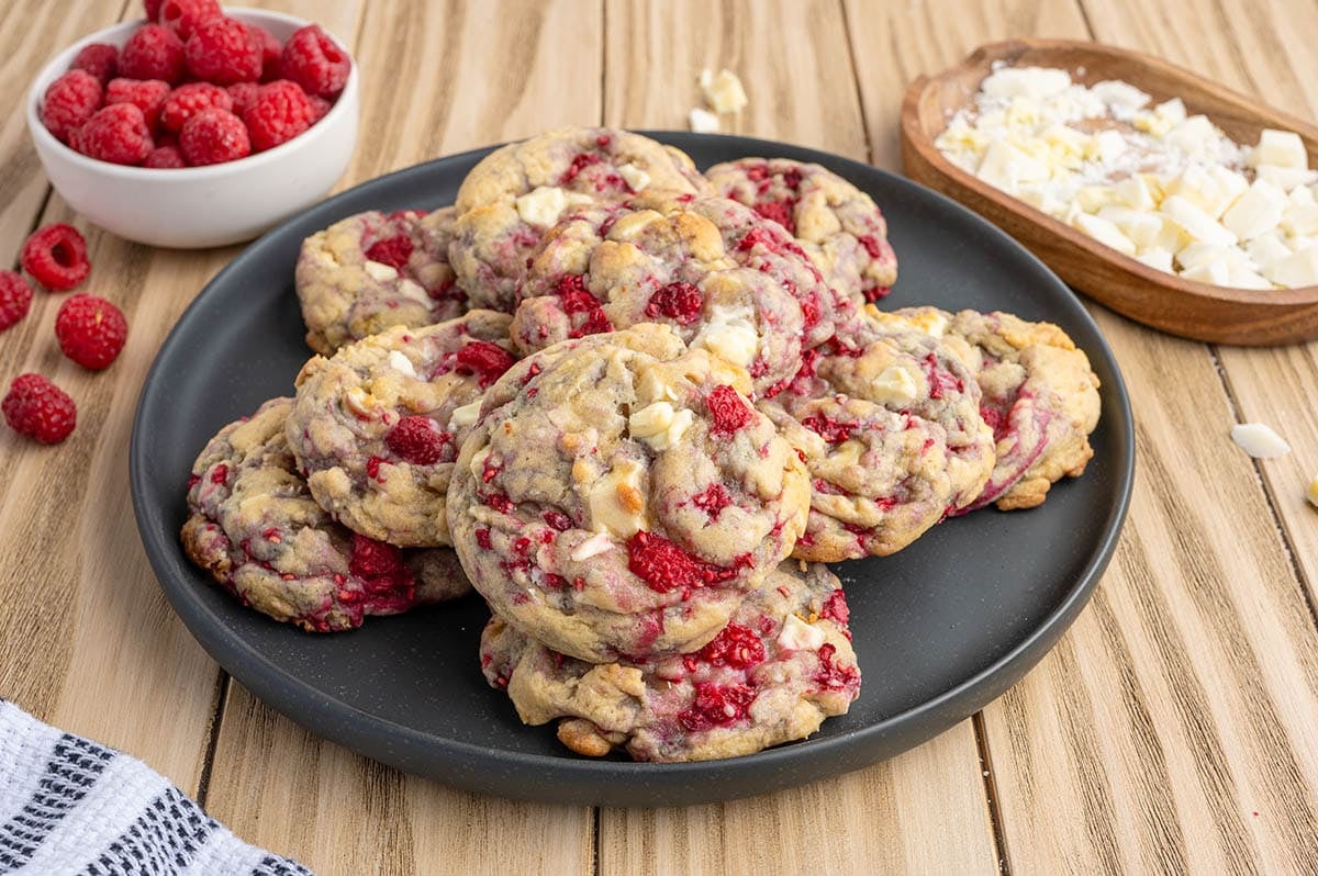 Stack of white chocolate raspberry cookies on a black plate with fresh raspberries and white chocolate chunks nearby.