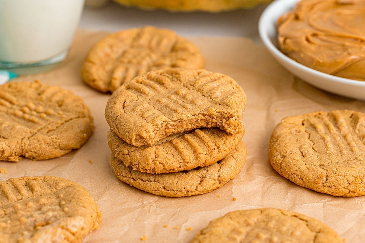 Golden peanut butter cookies stacked on parchment paper, one with a bite, milk glass and peanut butter bowl nearby.