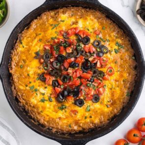 Cast iron skillet of cheesy taco dip topped with melted cheese, diced tomatoes, black olives, and cilantro.