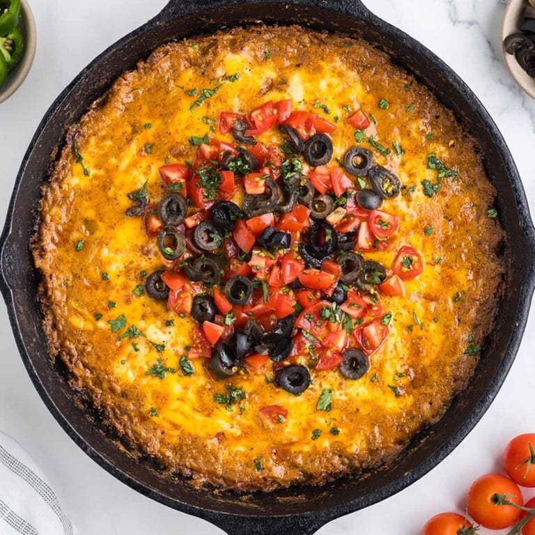 Cast iron skillet of cheesy taco dip topped with melted cheese, diced tomatoes, black olives, and cilantro.